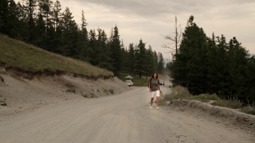 The girl is hitchhiking alone. He walks along the road with a paper map and tries to stop passing cars. Beautiful landscape: dusty road, forest, mountains.