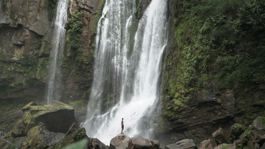 Man stands in front of large rain forest waterfall with leaves in foreground