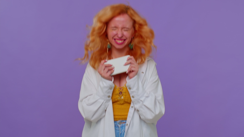 Worried funny addicted redhead girl enthusiastically playing racing video games on mobile phone. Young adult woman using smartphone gadget app with drive simulator isolayed on purple studio background