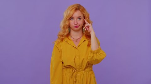 You are crazy, out of mind! Redhead girl in yellow dress pointing at camera and showing stupid gesture, blaming some idiot for insane plan. Young woman posing isolated on purple studio wall background