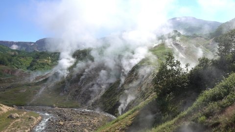A Unique Valley Of Geysers. Kamchatka.  Steam from hot springs rises into the blue sky above the mountain slopes. The river flows along a rocky bed. Green vegetation around