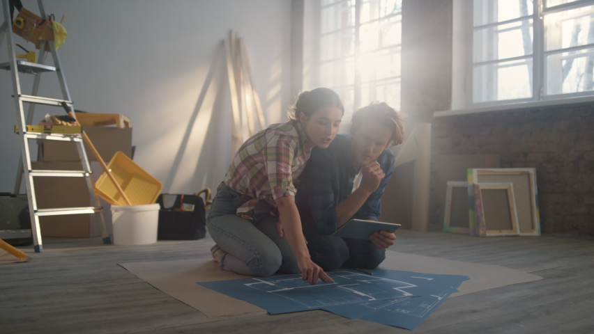 Serious woman explaining to man house renovation idea over blueprints. Closeup construction tools on blurred background. Focused couple discussing home repair scheme on floor in new apartment.