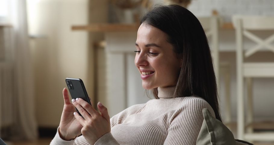 Relaxed happy young beautiful woman holding cellphone in hands, enjoying web surfing useful information, communicating distantly in social networks, playing mobile games or shopping in internet store. Royalty-Free Stock Footage #1077576581