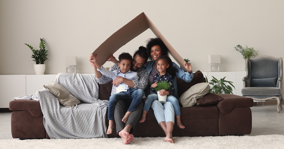 Happy emotional young african american family couple with little kids sitting on cozy sofa under carton roof, celebrating moving into new renovated apartment or house, relocation day concept. Royalty-Free Stock Footage #1077576611