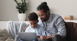 Curious african american small cute kid boy learning using computer software applications with caring father, sitting together on sofa. Addicted to modern tech happy biracial family playing on laptop.