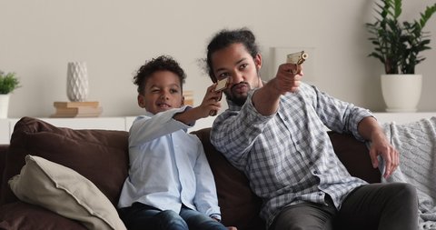 Carefree little african american preschool baby boy playing toy pistol, imagining shooting with loving young biracial father, sitting together on comfortable couch, happy family entertaining pastime.