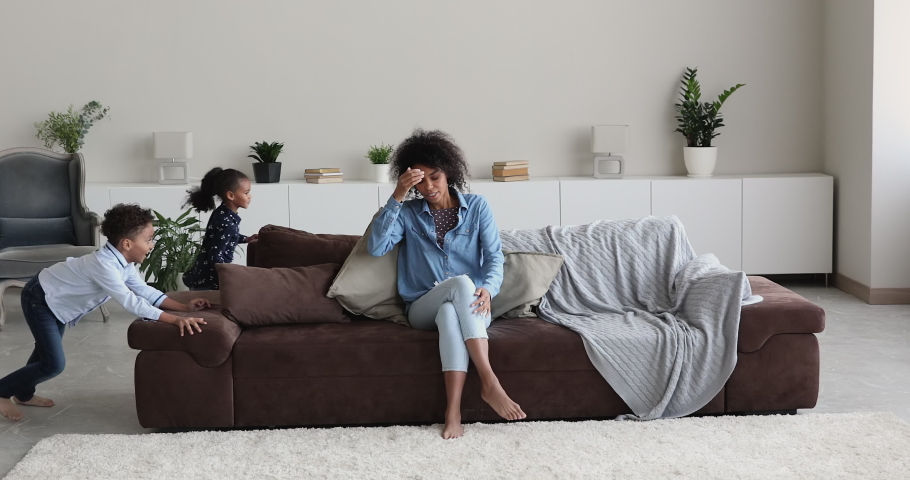 Stressed young african american mother sitting on couch, feeling tired of active noisy loud small children siblings playing running around in modern living room, upbringing difficulties concept. Royalty-Free Stock Footage #1077576665