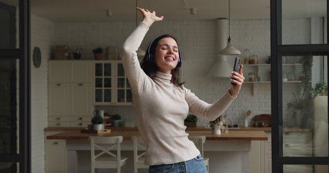 Happy sincere young woman holding cellphone in hands, choosing disco music from playlist, dancing to favorite audio in modern apartment. Candid female blogger recording streaming video on smartphone.