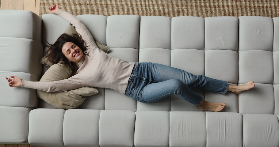 Top above view happy young beautiful smiling woman falling on huge comfortable sofa, raising arms stretching back muscles, enjoying carefree peaceful weekend relaxation time alone in living room.
