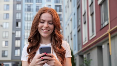Portrait of happy red hair beautiful woman with freckles typing by mobile phone outdoors. Cheerful girl walking with smartphone in urban street background. Smiling lady holding cellphone in hands.