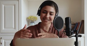 Indian woman in headphones sit indoor in front of microphone, look at laptop screen record podcast, talk to internet audience. On-air, online show broadcasting, livestream, host streaming live concept