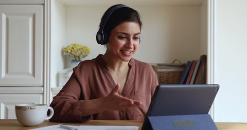 Happy young Indian woman in headphones start videoconference on digital tablet by business or study sit in homeoffice room. Video call event, apps, on-line tutor start virtual meeting class concept