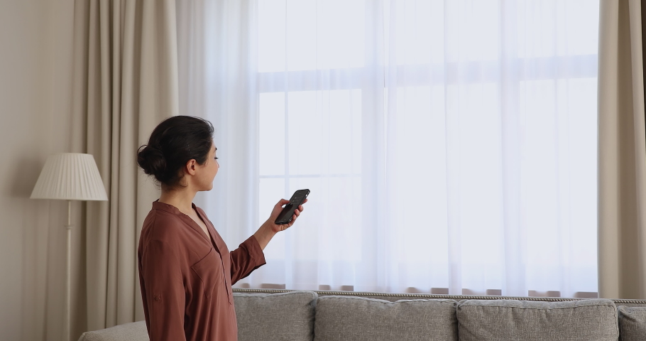 Young Indian woman close motorized window roller blinds in living room, holds cellphone use smart home automation mobile app, easy to control, contemporary house happy owner, modern tech usage concept | Shutterstock HD Video #1077583895