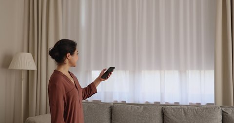 Young Indian woman close motorized window roller blinds in living room, holds cellphone use smart home automation mobile app, easy to control, contemporary house happy owner, modern tech usage concept