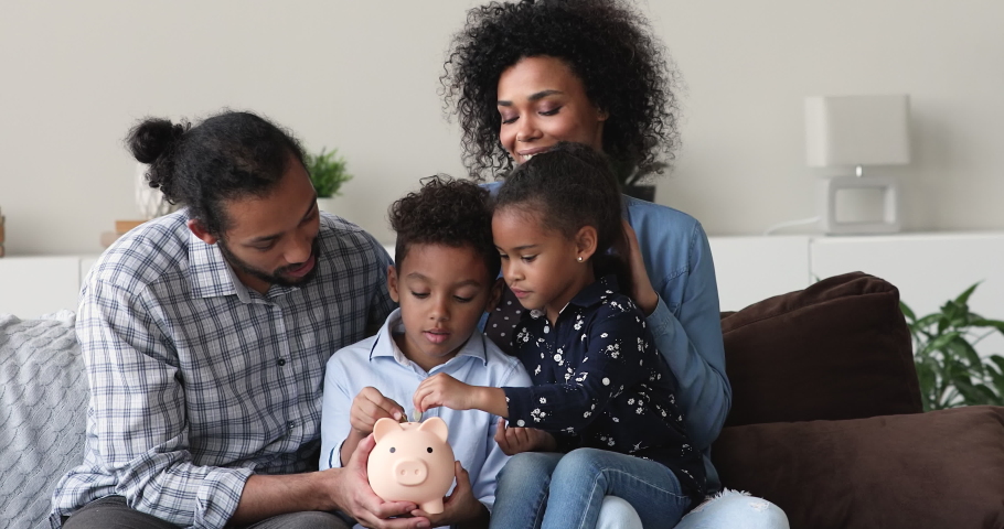 Smiling young african american couple parents holding on knees interested little children, teaching financial literacy at home. Curious small kids sibling learning saving money or budget management. Royalty-Free Stock Footage #1077583904