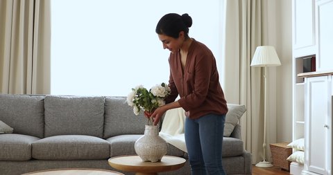 Young Indian ethnicity woman standing in modern cozy living room puts in vase bunch of beautiful fresh natural white wild roses. Happy housewife do routine housework, create cosiness at home concept