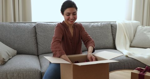 Indian woman sit on sofa unpack parcel with heap of boxes ordered for sell own handmade craft items, got gift from family, enjoy ordered ecommerce goods. Express delivery services, life event concept