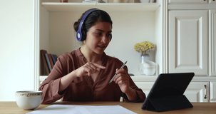 Indian woman in headphones sitting in kitchen using digital tablet talking by video call application, client and counsellor communicating remotely, working from home, videocall virtual event concept