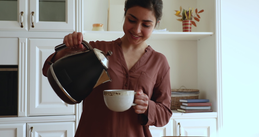 Young Indian woman standing in kitchen holding electric teapot pouring warm boiled water in ceramic cup preparing morning beverage for family breakfast. Healthy life habits, tea-time pleasure concept Royalty-Free Stock Footage #1077584009