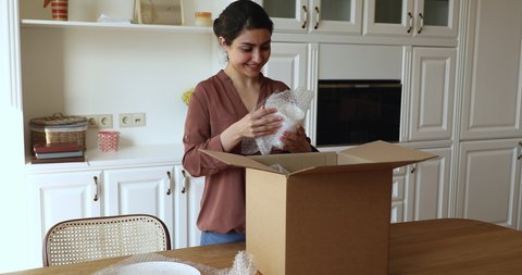 Indian woman standing in kitchen unpack parcel box with ordered fragile dishware crockery items wrapped in bubble wrap smiles feels satisfied by safe transport and e-commerce retail services concept