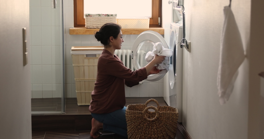 Indian housewife young woman do housework, busy in regular housekeeping work put bath towels into washing machine, turns on modern washer-dryer appliance in cozy laundry room. Household chores concept | Shutterstock HD Video #1077590324