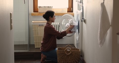Indian housewife young woman do housework, busy in regular housekeeping work put bath towels into washing machine, turns on modern washer-dryer appliance in cozy laundry room. Household chores concept
