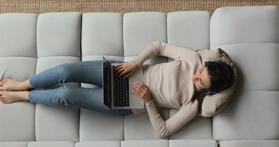 Overhead above top view relaxed young woman in wireless headphones holding web camera conversation using video call computer application, communicating distantly with friends resting on cozy sofa. Royalty-Free Stock Footage #1077590333