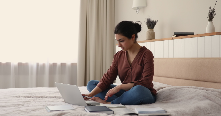 Indian woman sit on bed studying using laptop, writing information taken on internet, working on essay topic, makes research prepare for exams. Self-education, modern tech, e-learning remotely concept Royalty-Free Stock Footage #1077590345