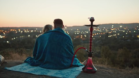 a guy and a girl in the mountains look at the sunset, wrapped in a blanket, they smoke a hookah, sit in an embrace. look at the night lights of the city. rest within the city.