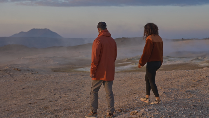 Slow motion panning shot of tourists, both clothed in orange coats, walking at a geothermal field in Iceland, with thin layer of smoke coming from below, in the backdrop of majestic pale blue skies