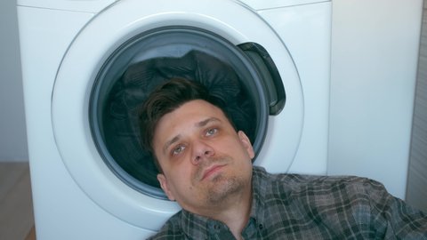 Tired man is waiting the washing machine with grey bedspread inside it, doing his laundry at home or laundry room. Everyday life, housework and household of young man.