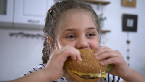 Little girl in fast food cafe eats burger. Portrait of hungry child Cute little girl eating burger. Hungry child in fast food cafe. Close-up girl eating burger. Hungry child in cafe. Lovely girl