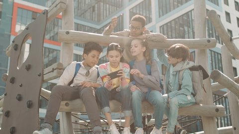 Low-angle slowmo shot of group of cheerful multiethnic school boys and girls playing games on smartphone at playground