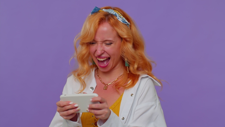 Worried funny addicted redhead girl enthusiastically playing racing video games on mobile phone. Young adult woman using smartphone gadget app with drive simulator isolayed on purple studio background