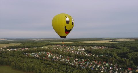 Yellow funny hot air balloon in the shape of a smile flight above flat field and forest landscapes at summer sunny sunset - Aerial drone wide view