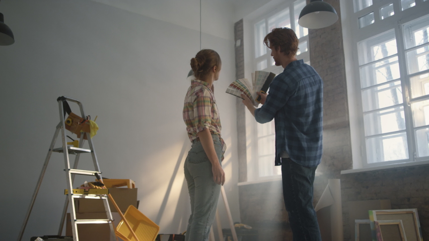 Back view of happy family choosing color for walls on window background. Handsome man holding color palette in hands for room decorating. Young couple staying among construction tools indoors. | Shutterstock HD Video #1077612926