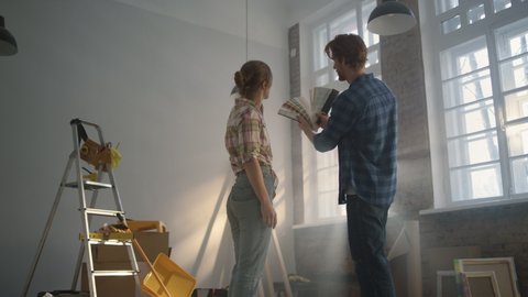 Back view of happy family choosing color for walls on window background. Handsome man holding color palette in hands for room decorating. Young couple staying among construction tools indoors.