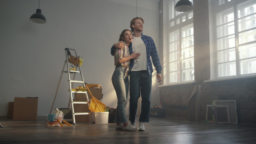 Cheerful couple admiring results of house renovation indoors. Amazed woman and man turning around in new apartment with tools for home repair. Happy family hugging indoors.