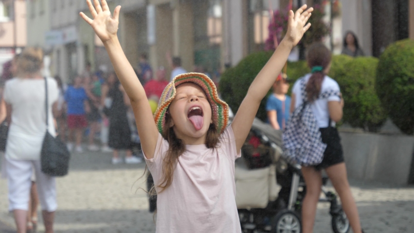 Funny little kid girl shows tongue catches drops under water mist cooling fog system on hot day in a city