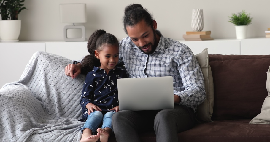 Caring young happy african american father teaching little cute kid daughter using computer software apps, resting together on comfortable sofa in modern living room, modern technology addiction. Royalty-Free Stock Footage #1077618455