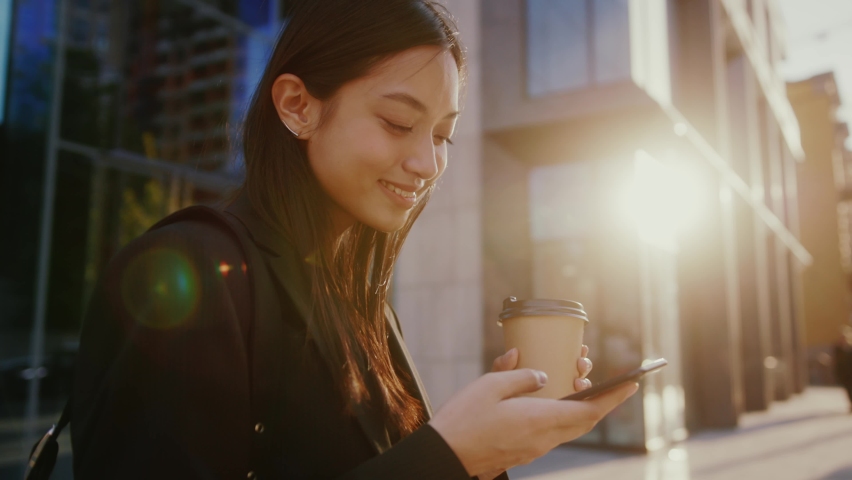 Inspired happy young asian mixed-race beautiful woman using smartphone, surfing internet on phone, enjoying morning sunshine outdoor. City. Modern people. Royalty-Free Stock Footage #1077619085