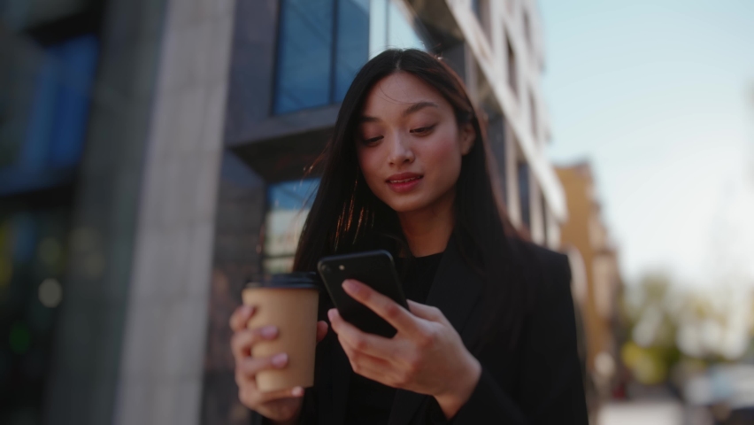 Joyful young asian business girl using mobile phone networking with online friends typing messages drinking coffee outside. Beautiful people. City life. Royalty-Free Stock Footage #1077619097