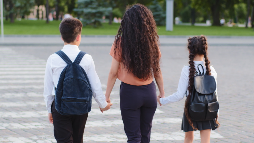 Slow motion back view mother woman with two children pupils schoolchildren holding hands of daughter and son little girl and small boy going stepping on pedestrian crossing walking along road in city Royalty-Free Stock Footage #1077619484