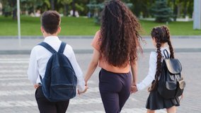 Slow motion back view mother woman with two children pupils schoolchildren holding hands of daughter and son little girl and small boy going stepping on pedestrian crossing walking along road in city - Powered by Shutterstock - Get 15% off with code: PIKWIZARD15