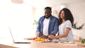 Joyful mixed race couple enjoying cooking with a video tutorial, happy African boyfriend and biracial girlfriend preparing meal in front of the laptop, a guy is biting slice of tomato and smiles