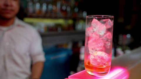 Professional Asian barman pouring liquor into cocktail glass with ice cube on counter bar serve in nightclub. Male mixologist bartender preparing alcohol cocktail drink with liquor shelf background