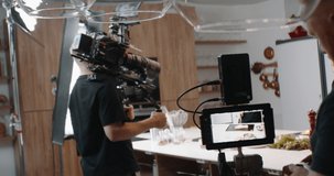BTS of a cooking TV show - celebrity host cooking a dish, talking into camera. Camera operator and focus puller in the frame