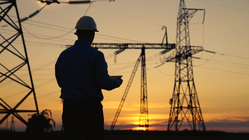 Silhouette Power engineer in white helmet checks power line using computer on tablet, remote control of power system. High voltage electrical lines at sunset. Distribution and supply of electricity