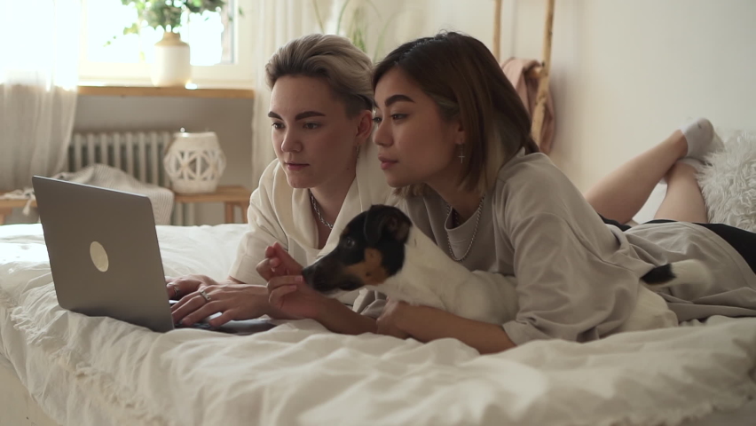 Lesbian couple of women using laptop and lying on bed in home bedroom spbd.