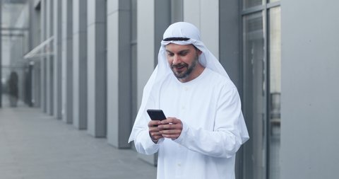 Waist up portrait view of the arabian businessman wearing kandora looking at his mobile phone and rejoicing while feeling happy of the successful business deal